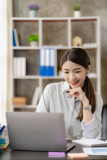 Sorrindo jovem empresária asiática trabalhando em um computador portátil em sua mesa e calculando despesas financeiras. gráfico folha de dados de papel imagem vertical - Foto, Imagem