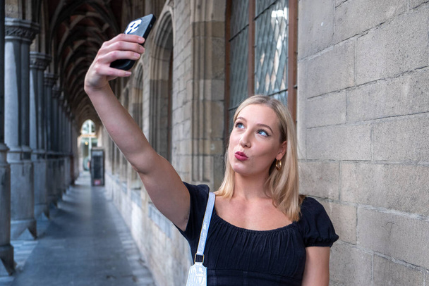 Πορτρέτο του υπέροχο λευκό γυναικείο μοντέλο με φωτεινό μακιγιάζ εκφράζοντας ενέργεια σε καλή μέρα στην Ευρώπη. Υπέροχη ξανθιά γυναίκα με κομψά ρούχα που κάνει selfie, ενώ το περπάτημα παρελθόν παλιό κτίριο. Υψηλή ποιότητα - Φωτογραφία, εικόνα