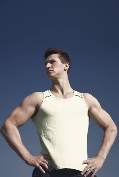 Athlete in green tshirt on sunny day. Man bodybuilder posing on blue sky. Workout and training. Sport and healthy lifestyle concept. Fitness model with muscular hands, biceps, triceps, strong muscles. - Фото, зображення
