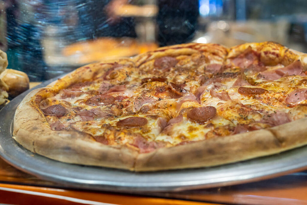 Peperoni und Speck-Pizza in Scheiben geschnitten und auf einer Theke hinter dem Glas einer Glasvase ausgestellt. - Foto, Bild