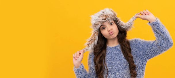 lachend kind in trui en oorflap hoed op gele achtergrond, wintertijd. Banner van kind meisje in de winter hoed, studio poster header met kopieerruimte - Foto, afbeelding