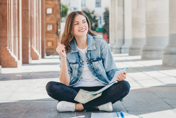 Studentka sedí naproti univerzitě a usmívá se. Roztomilá studentka drží pensílie, složky, zápisníky a směje se. Dívka učí lekce - Fotografie, Obrázek