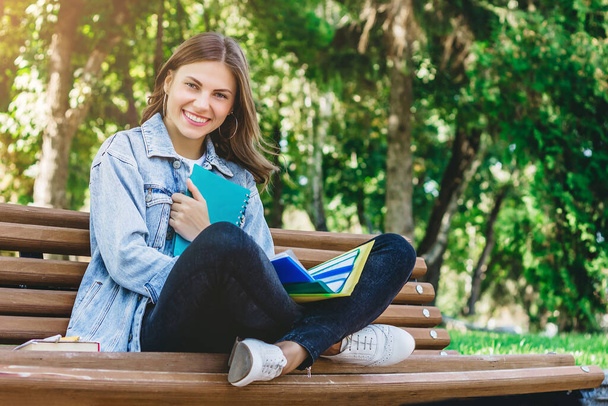 Νεαρή μαθήτρια κάθεται σε ένα παγκάκι στο πάρκο και κρατά βιβλία, σημειωματάρια και φακέλους. Το κορίτσι διδάσκει στο πάρκο.. - Φωτογραφία, εικόνα