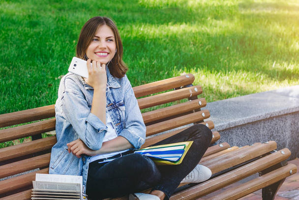 Die junge Studentin sitzt auf einer Bank im Park und hält Bücher, Notizbücher und Mobiltelefone in der Hand. Mädchen erteilt Unterricht im Park. - Foto, Bild
