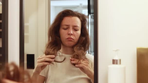 Nešťastná mladá brunetka v koupelně zrcadlo nalezení poškozené vlasy, pocit nespokojenosti s vyschlostí nebo roztřepených koncích, myšlení na lázeňské profesionální procedury.  - Záběry, video