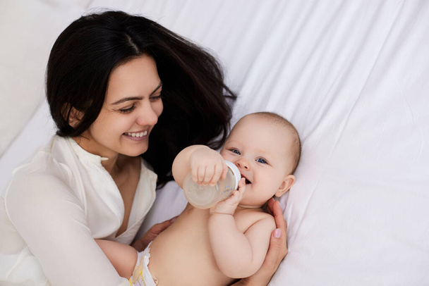 happy mother feeding her newborn baby on the bed at home. mom holding a bottle of water - Foto, imagen