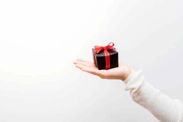 赤いリボンで包まれた小さな贈り物を持つ女性の手のショットを閉じます。コピースペース、バナーと休日プレゼントのコンセプト。小さな箱に焦点を当て、フィールドの浅い深さ. - 写真・画像