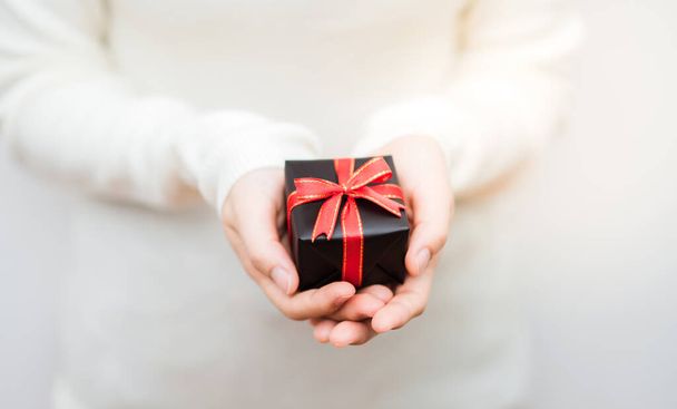 赤いリボンで包まれた小さな贈り物を持つ女性の手のショットを閉じます。コピースペース、バナーと休日プレゼントのコンセプト。小さな箱に焦点を当て、フィールドの浅い深さ. - 写真・画像