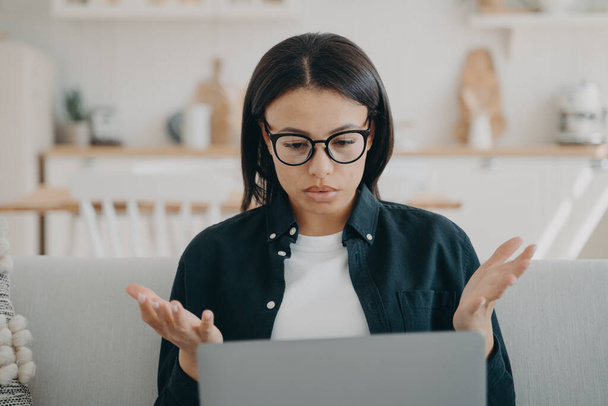 Puzzled woman working at laptop spreading hands, reading email with bad news at home. Frustrated female freelancer in glasses looks at screen, shocked by problems in work, error on computer. - Photo, Image