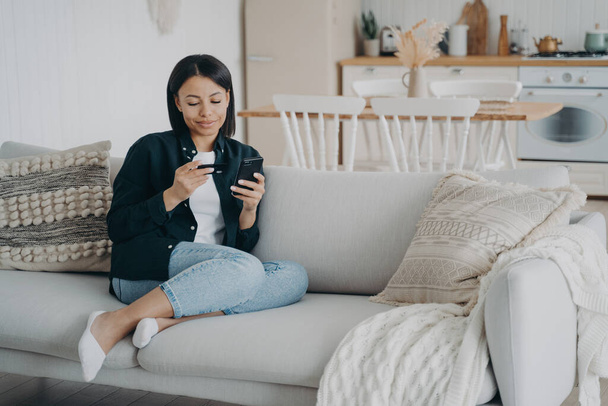 Woman holding credit card, phone makes cashless payment, shopping on internet, sitting on cozy sofa at home. Female checking bank account, using mobile app. E-commerce, online banking service. - Photo, image