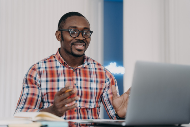 Sonriendo desconcertado hombre afroamericano leyendo correo electrónico con noticias trabajando en el ordenador portátil, extendiendo sus manos. Estudiante negro mirando la pantalla de la computadora decidiendo tareas de estudio, aprendiendo en línea. - Foto, imagen