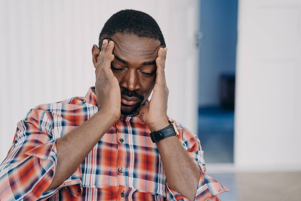 Yorgun, üzgün Afrikalı Amerikalı bir adam başı ellerindeyken baş ağrısından muzdarip. Çok çalışan, hayal kırıklığına uğramış, migren ağrısı çeken siyah bir adam. İş stresi, tükenme, yaşam krizi, başarısızlık.. - Fotoğraf, Görsel