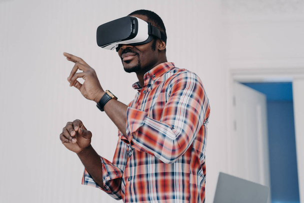 Homme afro-américain portant des lunettes VR touchant des objets virtuels par les doigts, interagissant avec le cyberespace. Homme noir moderne en réalité virtuelle lunettes de jeu vidéo en ligne. Concept de haute technologie. - Photo, image