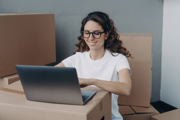 Dizüstü bilgisayarda çalışan, karton kutularla çevrili gülümseyen İspanyol kadın müşteri siparişleri hazırlıyor, gönderilmek üzere çevrimiçi mağaza paketleri hazırlıyor. Teslimat hizmeti, e-ticaret konsepti. - Fotoğraf, Görsel