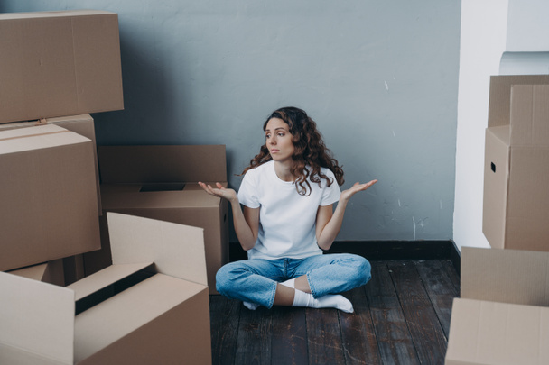 Puzzled young girl shrugging, sitting on the floor with cardboard boxes, having problem with moving to new home. Hard choice, uncertainty, dilemma and relocation. Advertising of moving company service - Фото, изображение