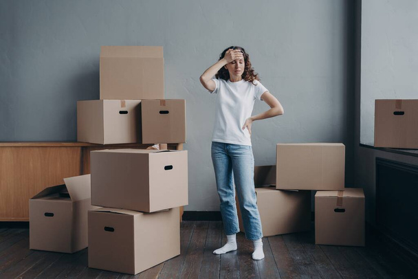 Tired upset woman holding her head standing in empty room near heap of cardboard boxes feel unmotivated to pack things. Financial problem, debt, eviction, divorce, hard relocation day concept. - Photo, Image