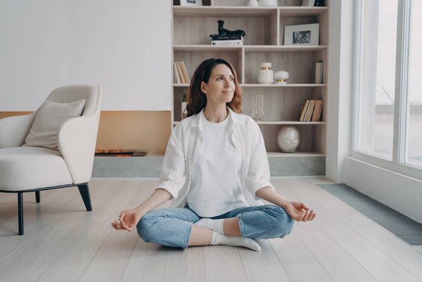 Mujer hispana pacífica practicando yoga sentada en padmasana, pose de loto en el suelo en la sala de estar. Calma mujer meditando, haciendo ejercicios de respiración en casa. Alivio del estrés, concepto de estilo de vida saludable. - Foto, Imagen