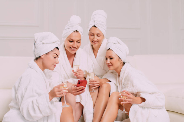 A group of women spend leisure time. Four Young attractive women in bathrobes look at the smartphone screen and smile. - Photo, image