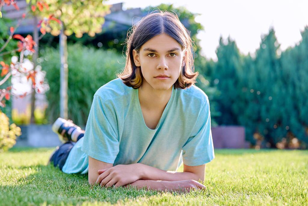 Portrait of a serious calm young guy looking at the camera, lying on the grass. Handsome teenage male resting posing on the lawn in the backyard, in the park - Zdjęcie, obraz