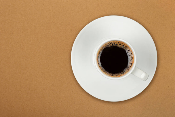 Close up one white cup full of black coffee on saucer over brown paper parchment background, elevated top view, directly above - Photo, Image