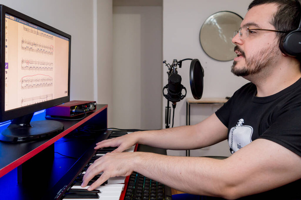 jovem caucasiano latino com barba, óculos e fones de ouvido, sentado tocando piano e cantando, estudando partituras digitais com o computador em casa - Foto, Imagem
