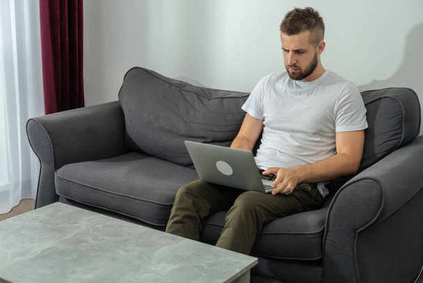 Nuori hymyilevä mies rentoutuu työskennellessään kannettavan tietokoneen ja videoneuvottelun kanssa kotona. Nuori luova mies katselee näytön viestin älypuhelimella.Työ kotoa - Valokuva, kuva