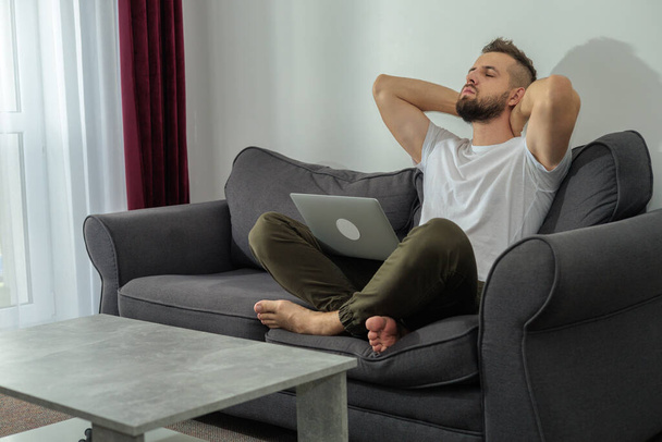 Fiatal mosolygós férfi pihentető dolgozik laptop és videó konferencia otthon. Fiatal kreatív férfi néz képernyő üzenet segítségével okostelefon.Munka otthonról - Fotó, kép
