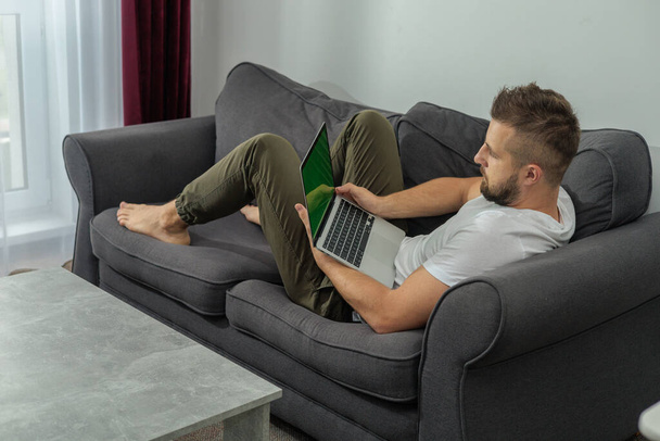 Jonge glimlachende man ontspannen werken met laptop en video conferentie thuis. Jonge creatieve man kijkt naar het scherm bericht met behulp van smartphone.Work van thuis uit - Foto, afbeelding