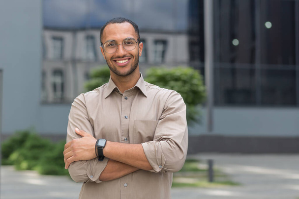 Portrait of young hispanic student man smiling and looking at camera near university campus with arms crossed wearing glasses and shirt. - Valokuva, kuva