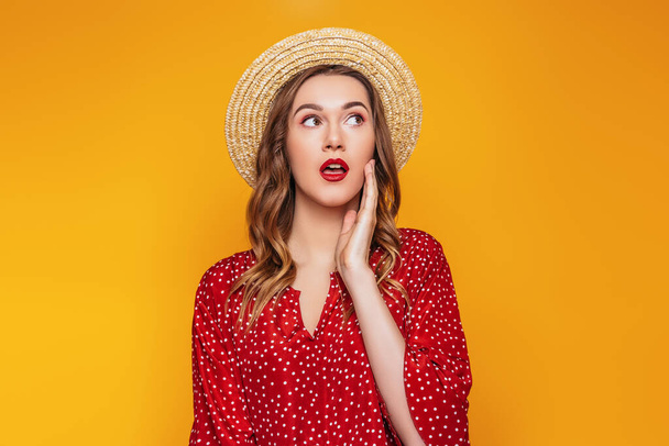 Chica joven sorprendida en un sombrero de paja, en un vestido rojo con un lápiz labial rojo mira hacia otro lado en el espacio de copia, retrato de una mujer joven conmocionada en ropa de verano sobre un fondo naranja, concepto de venta - Foto, Imagen