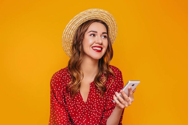 Młoda dziewczyna w czerwonej letniej sukience, słomkowym kapeluszu z czerwonymi ustami trzyma telefon komórkowy, smartfona w rękach, śmieje się, uśmiecha i patrzy na przestrzeń odizolowaną na żółtym, pomarańczowym tle - Zdjęcie, obraz