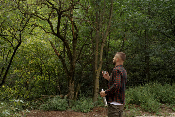 Bir adam ormanda yürüyor, dinleniyor ve termostan su içiyor. - Fotoğraf, Görsel