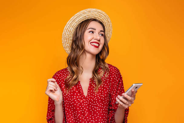 Young stylish girl in a red vintage dress, a straw hat, with red lipstick smiles, laughs, listens to music on the mobile phone and looks at copy space isolated on a yellow, an orange background - Foto, Imagem