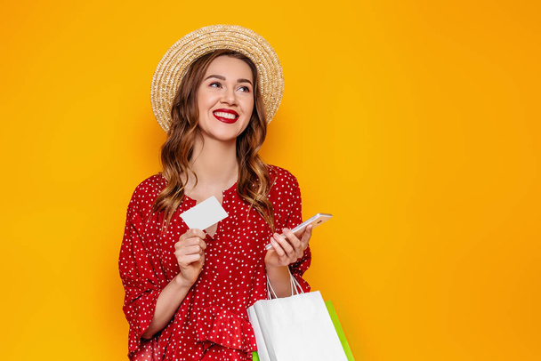 Kaunis nuori nainen punaisessa kesämekossa, olkihattu pitää matkapuhelinta ja luottokorttia käsissään eristettynä keltaisella taustalla, kopiotila, mallinnus, web banneri. Tyttö tekee verkko-ostoksia - Valokuva, kuva
