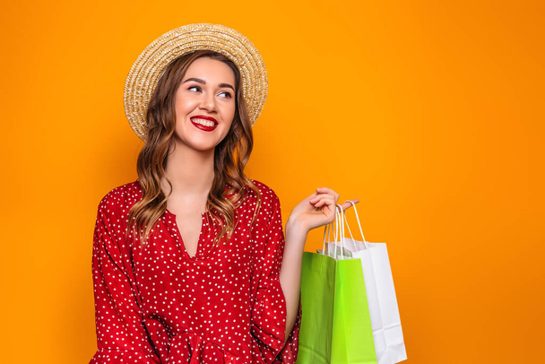 Hermosa joven con un vestido rojo de verano, sombrero de paja sostiene una bolsa de compras en sus manos aisladas sobre un fondo naranja, espacio de copia. Chica con regalos de compras mira el espacio de copia - Foto, Imagen