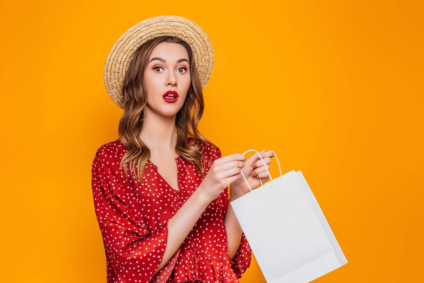 Portrait of a shocked surprised girl dressed in a red dress and straw hat with red lips holding shopping bags and looking at camera isolated over orange background - Valokuva, kuva
