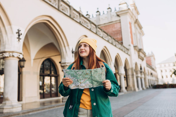 Aantrekkelijke jonge vrouwelijke toerist verkent nieuwe stad. Een roodharige meid met een papieren kaart op het marktplein in Krakau. Reizen door Europa - Foto, afbeelding