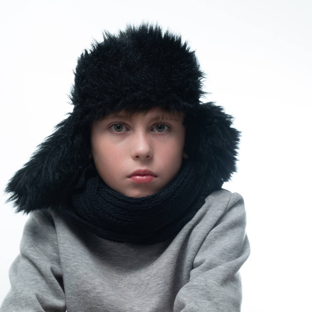 Portrait of a child in a winter hat with earflaps and a knitted scarf on a white background, black winter clothes. - Photo, Image