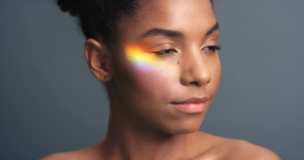 Rainbow, prism and light on face of black woman for glow, beauty or LGBT pride against a grey background in studio. Holographic, flare and reflection on young female for art, creative or skincare. - Filmagem, Vídeo