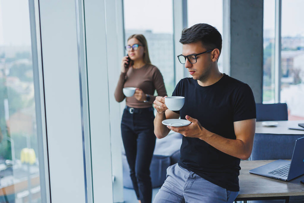 Businessman holds a cup of coffee in his hand. Multitasking concept. Busy freelancer in glasses drinking tea and talking in a cafe. smart casual. - Foto, Bild