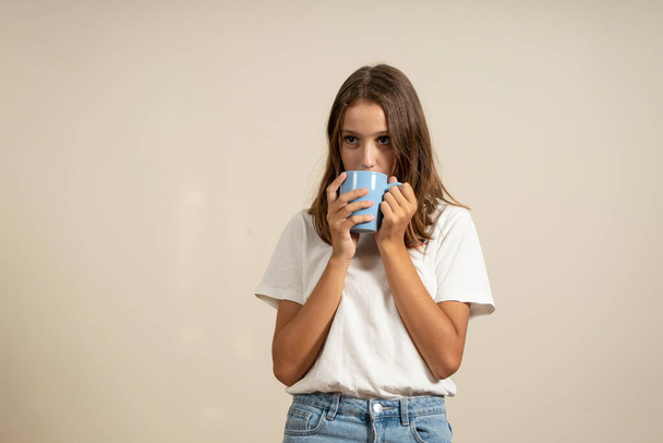 Gorgeous Hispanic teenage girl with brunette hair drinking a cup of hot chocolate isolated on beige studio background - 写真・画像
