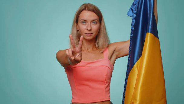 Žena držící ukrajinskou národní vlajku, ukazující znamení vítězství nad válkou, doufající v úspěch a vítězství, činící mírové gesto, usmívající se s laskavým optimistickým výrazem. Mladá dívka na modrém pozadí studia - Fotografie, Obrázek
