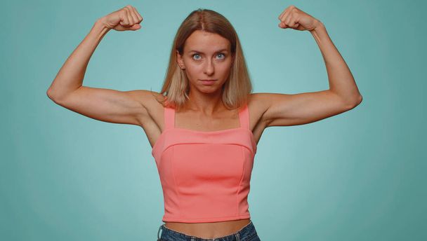Ik ben sterk en onafhankelijk. Vrouw toont biceps, kijkt zelfverzekerd, voelt kracht om te vechten voor rechten, energie om succes te winnen. jong volwassen meisje geïsoleerd op blauw studio muur achtergrond - Foto, afbeelding