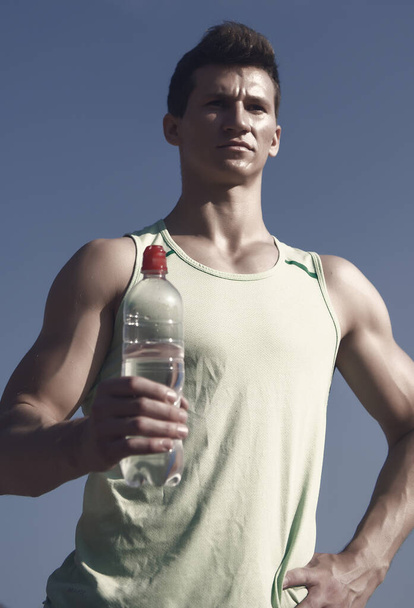 Man with water bottle in muscular hands. Athlete in green tshirt on blue sky. Bodybuilder with strong muscles biceps, triceps on sunny day. Sport and fitness concept. Healthy lifestyle and health. - Fotó, kép