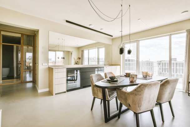 Современный минималистский дизайн интерьера однокомнатной квартиры с открытой белой кухней и обеденной зоной со столом и стульями, освещенными лампой в стиле лофт - Фото, изображение