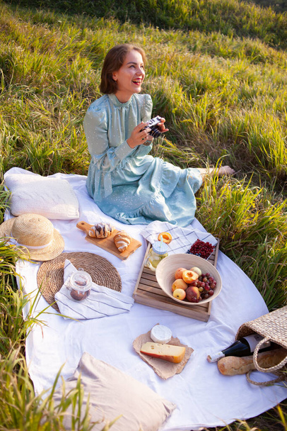 Laughing woman in a long dress with short hair sitting on a white blanket with fruits and pastries and looking at the pictures on camera. Concept of having picnic during summer holidays or weekends - Photo, Image