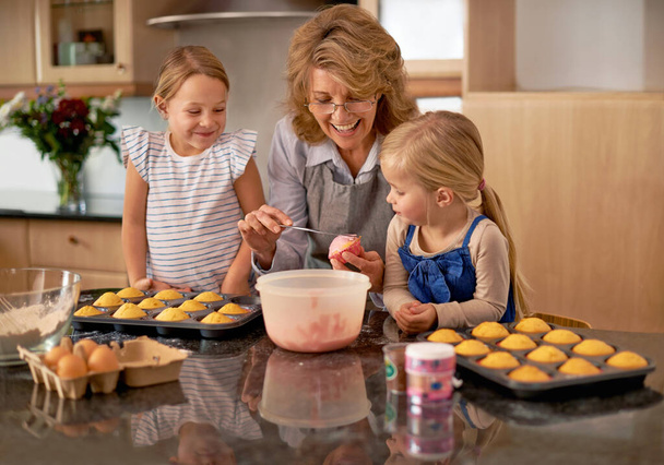 Haar kleinkinderen waardevolle keukenvaardigheden bijbrengen. Twee kleine meisjes bakken cupcakes met de hulp van hun oma thuis - Foto, afbeelding