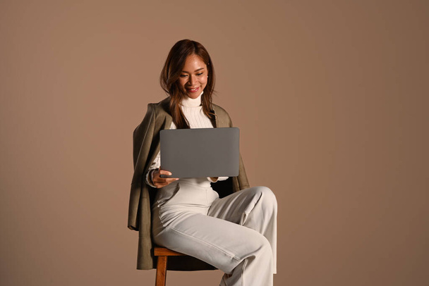 Bild einer lächelnden jungen Frau im Trenchcoat, die vor braunem Hintergrund sitzt und Laptop benutzt. Mensch und Technologie. - Foto, Bild