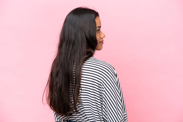Young Colombian woman isolated on pink background in back position and looking side - Photo, Image