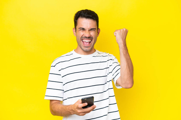 Joven hombre guapo sobre fondo amarillo aislado con teléfono en posición de victoria
 - Foto, Imagen